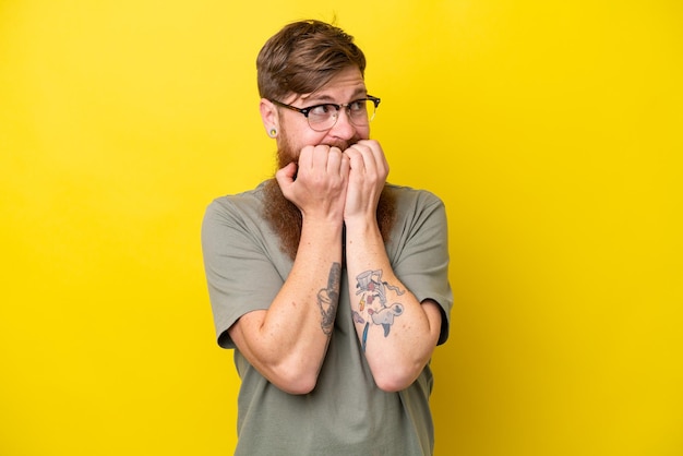 Redhead man with beard isolated on yellow background nervous and scared putting hands to mouth