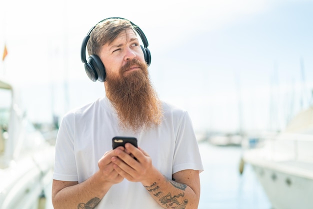 Redhead man with beard listening music with a mobile and thinking