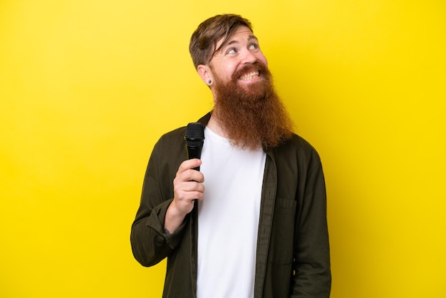 Redhead man with beard picking up a microphone isolated on yellow background looking up while smiling