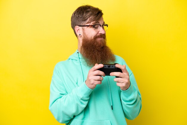 Redhead man with beard playing with a video game controller isolated on yellow background with happy expression