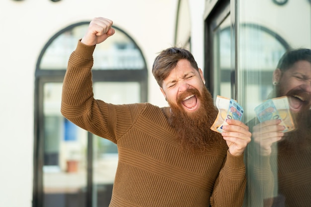 Photo redhead man with beard taking a lot of money and celebrating a victory