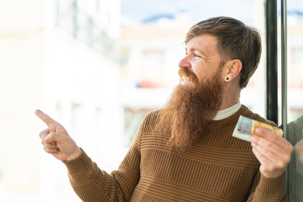 Photo redhead man with beard taking a lot of money at outdoors pointing to the side to present a product