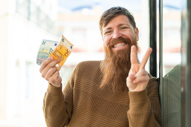 Redhead man with beard taking a lot of money at outdoors smiling and showing victory sign