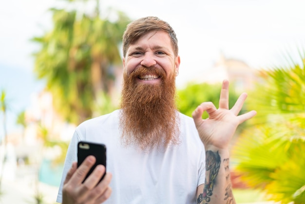 Redhead man with beard using mobile phone and doing OK sign