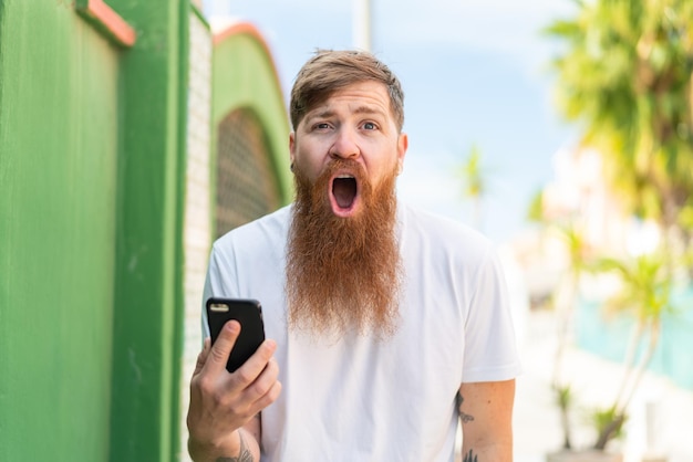 驚きとショックを受けた表情で屋外で携帯電話を使用してひげを持つ赤毛の男