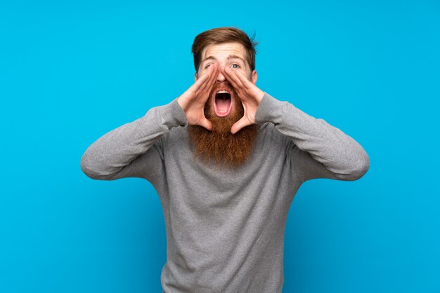 Redhead man with long beard over blue wall shouting and announcing something