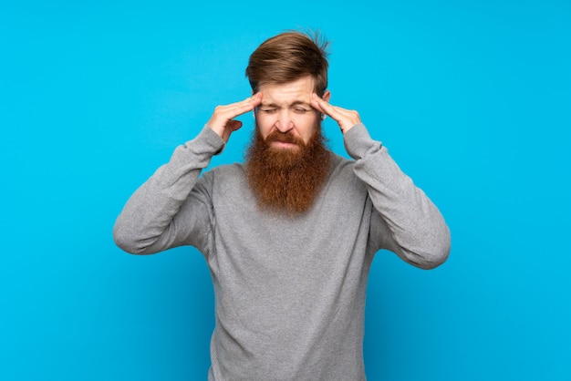 Redhead man with long beard over blue wall with headache