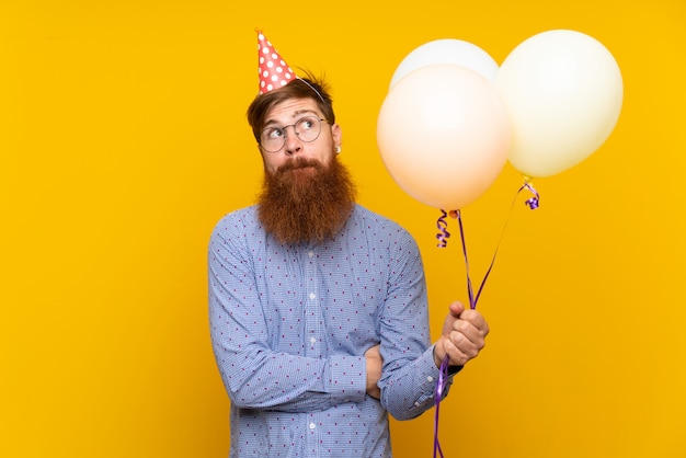 Redhead man with long beard holding balloons over isolated yellow wall making doubts gesture while lifting the shoulders