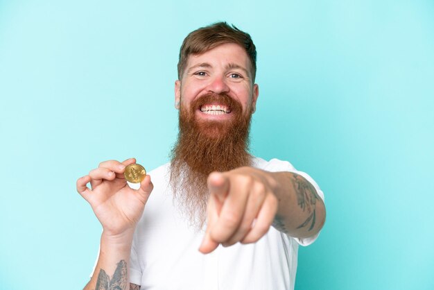 Redhead man with long beard holding a Bitcoin isolated on blue background points finger at you with a confident expression