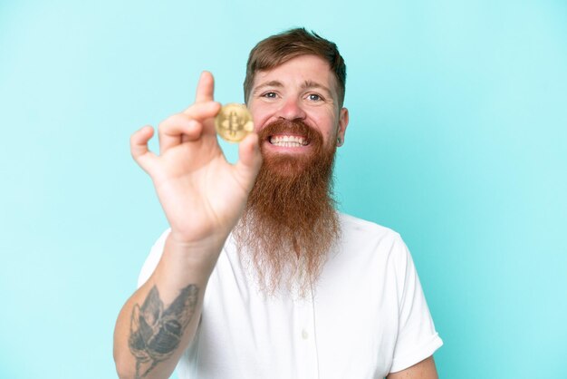 Redhead man with long beard holding a Bitcoin isolated on blue background with happy expression