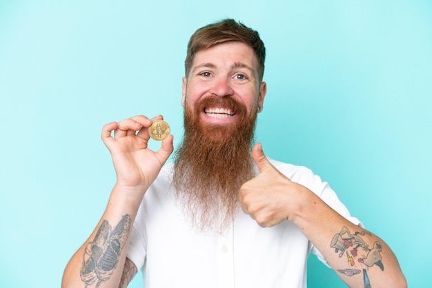 Redhead man with long beard holding a Bitcoin isolated on blue background with thumbs up because something good has happened
