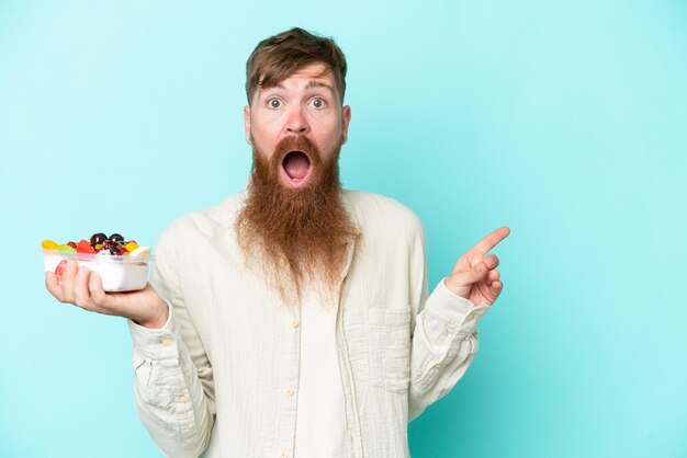 Redhead man with long beard holding a bowl of fruit isolated on blue background surprised and pointing finger to the side