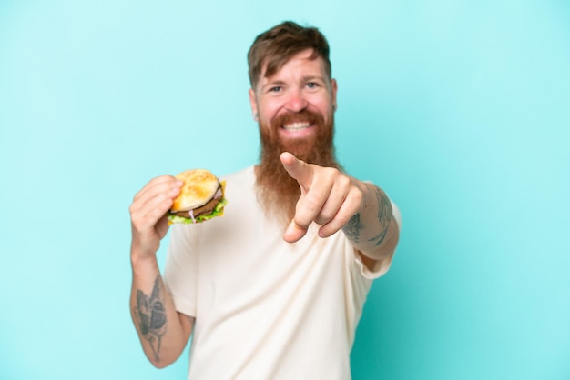 Redhead man with long beard holding a burger isolated on blue background points finger at you with a confident expression