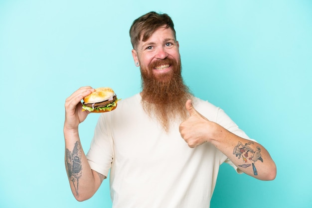 긴 수염을 가진 빨간 머리 남자는 좋은 일이 일어났기 때문에 엄지손가락을 치켜들고 파란색 배경에 격리된 햄버거를 들고 있습니다.