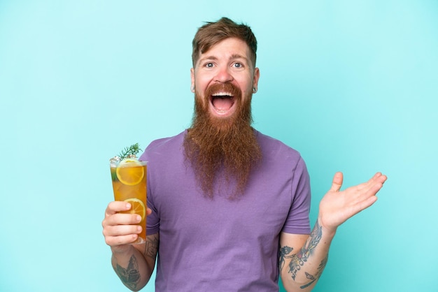 Redhead man with long beard holding a cocktail isolated on blue background with shocked facial expression