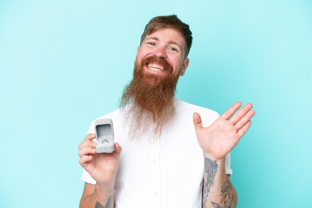 Redhead man with long beard holding a engagement ring isolated on blue background saluting with hand with happy expression