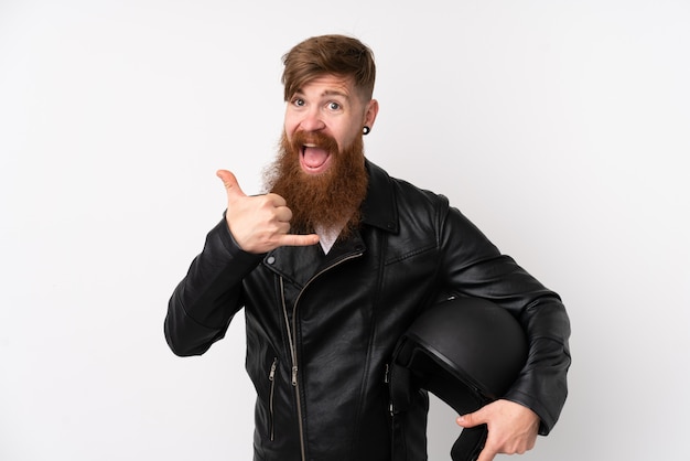 Redhead man with long beard holding a motorcycle helmet over isolated white wall making phone gesture