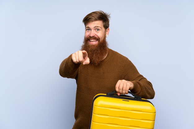 Redhead man with long beard holding a suitcase points finger at you with a confident expression