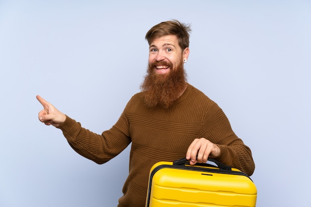 Redhead man with long beard holding a suitcase surprised and pointing finger to the side