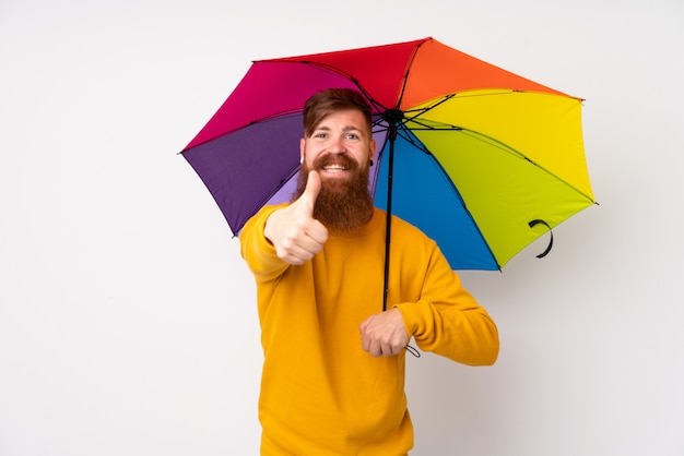 Redhead man with long beard holding an umbrella over isolated white wall giving a thumbs up gesture