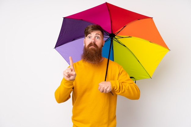 Redhead man with long beard holding an umbrella over isolated white wall pointing with the index finger a great idea