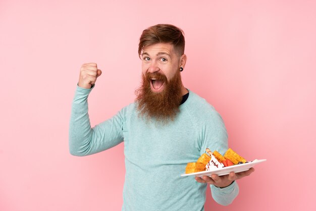 L'uomo di redhead con la barba lunga che tiene i waffles sopra la parete dentellare isolata che celebra una vittoria