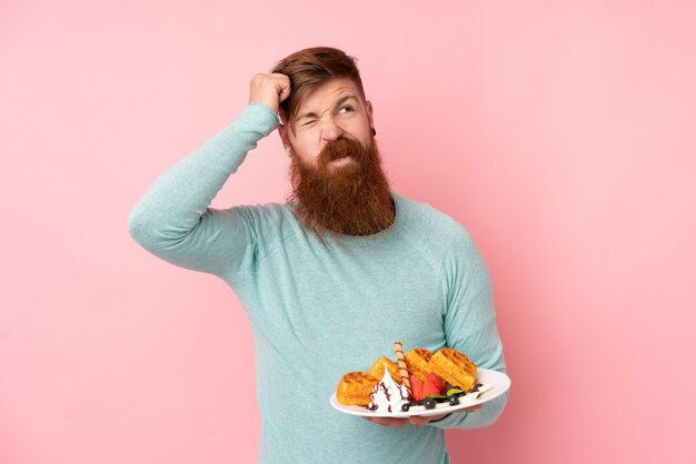 Photo redhead man with long beard holding waffles over isolated pink wall having doubts and with confuse face expression