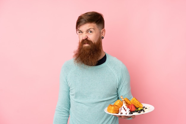 Redhead man with long beard holding waffles over isolated pink wall making doubts gesture while lifting the shoulders