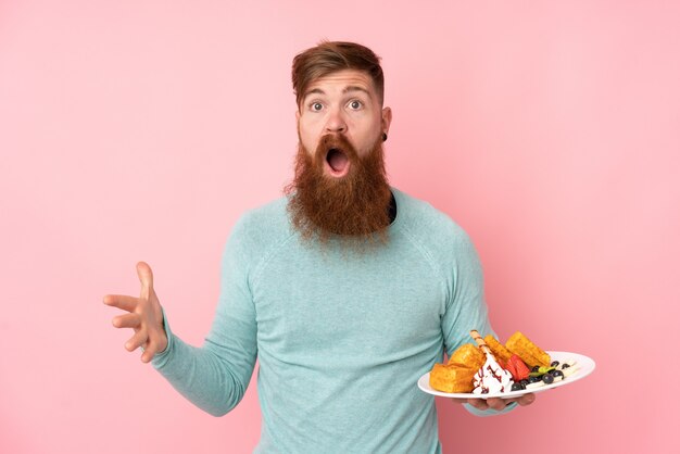 Redhead man with long beard holding waffles over isolated pink wall with shocked facial expression