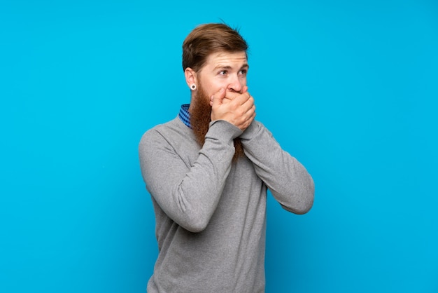 Photo redhead man with long beard over isolated blue covering mouth and looking to the side