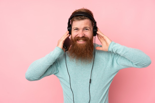 Redhead man with long beard over isolated pink wall listening music