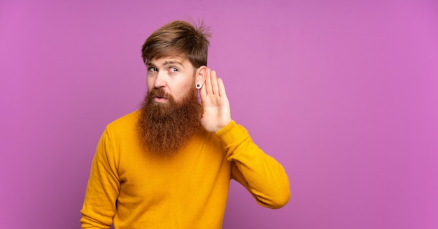 Redhead man with long beard over isolated purple listening to something by putting hand on the ear