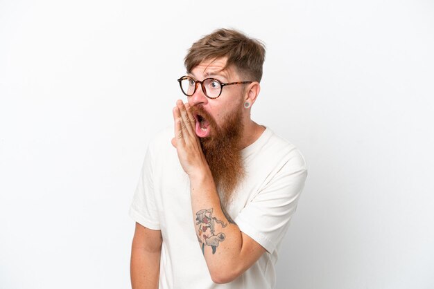 Redhead man with long beard isolated on white background whispering something with surprise gesture while looking to the side