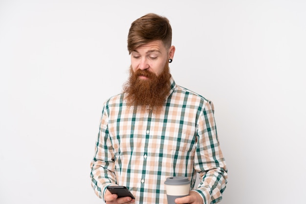 Рыжий мужчина с длинной бородой над изолированной белой стеной держит кофе на вынос и мобильный