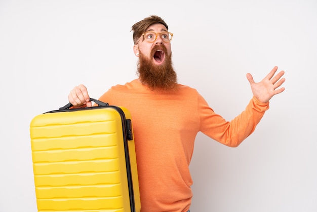 Redhead man with long beard over isolated white wall in vacation with travel suitcase and surprised