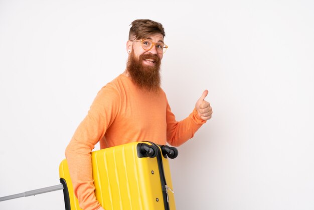 Redhead man with long beard over isolated white wall in vacation with travel suitcase and with thumb up