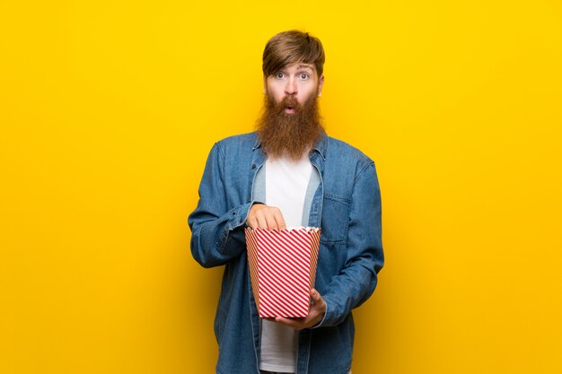 Redhead man with long beard over isolated yellow wall eating popcorns