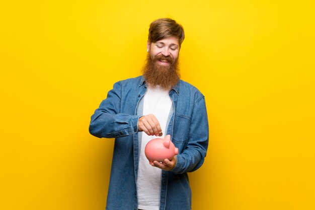 Uomo di redhead con barba lunga sopra isolato muro giallo in possesso di un grande salvadanaio