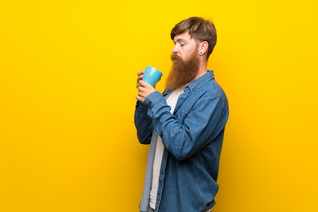 Redhead man with long beard over isolated yellow wall holding hot cup of coffee