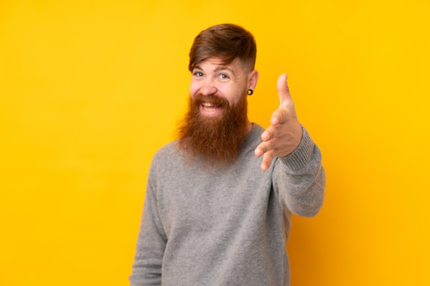 Redhead man with long beard over isolated yellow wall shaking hands for closing a good deal