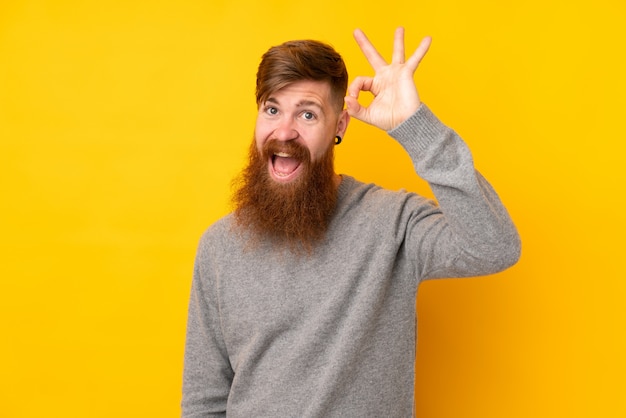 Redhead man with long beard over isolated yellow wall surprised and showing ok sign