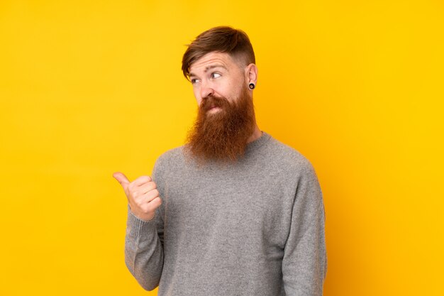 Redhead man with long beard over isolated yellow wall unhappy and pointing to the side