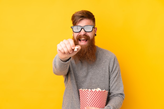 Redhead man with long beard over isolated yellow wall with 3d glasses and holding a big bucket of popcorns while pointing front