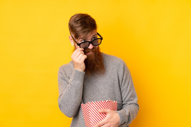 Redhead man with long beard over isolated yellow wall with 3d glasses and holding a big bucket of popcorns