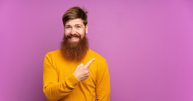 Redhead man with long beard over purple wall pointing to the side to present a product