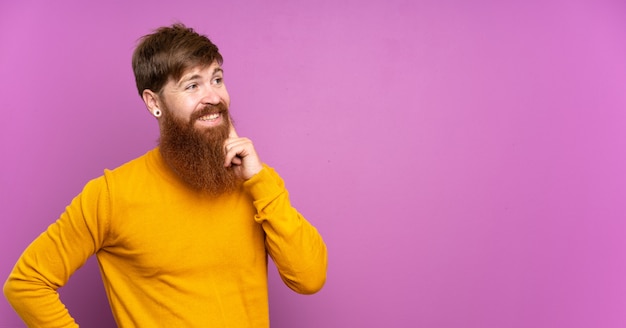 Uomo di redhead con barba lunga sul muro viola pensando un'idea mentre guardando in alto