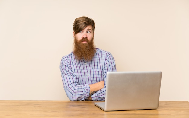 Redhead man with long beard in a table with a laptop thinking an idea