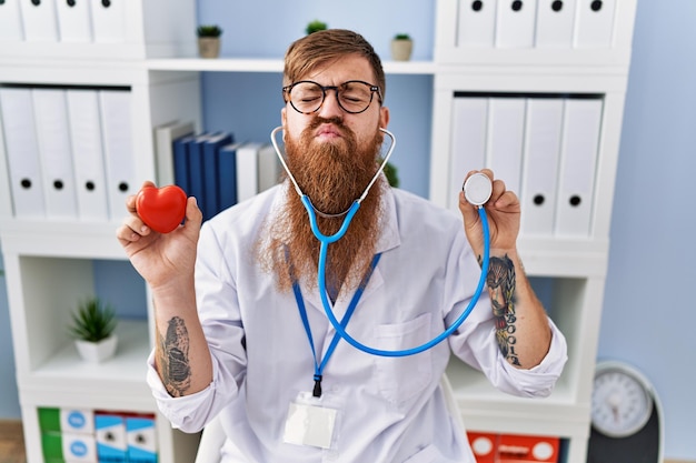 Redhead man with long beard wearing doctor uniform holding heart and stethoscope looking at the camera blowing a kiss being lovely and sexy love expression