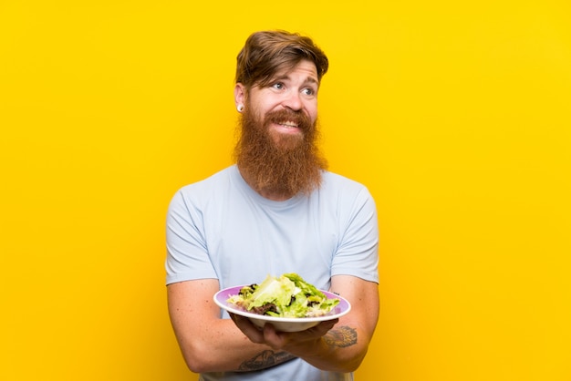 redhead man with long beard and with salad over isolated yellow wall looking up while smiling