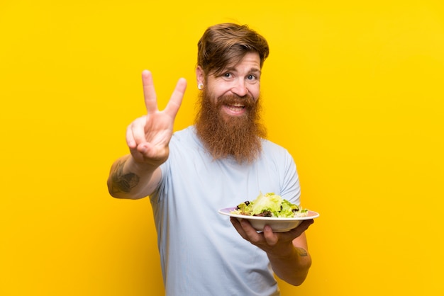 Redhead man with long beard and with salad over isolated yellow wall smiling and showing victory sign